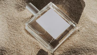 Ein Flakon liegt im Sand mit einer Muschel daneben.
