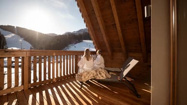 Zwei Frauen sitzen auf einem Holz-Balkon mit Bademänteln und trinken Wein. Der Ausblick ist eine Landschaft in den Bergen mit Schnee.