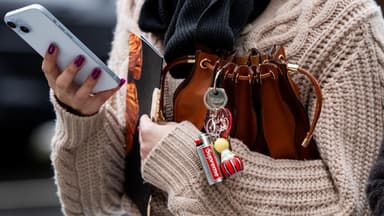 Frau mit einem Cardigan hält ihre Tasche unterm Arm und ein Handy in der Hand. An der Tasche ist ein Schlüsselanhänger.