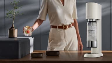 Eine Frau hält ein Glas mit Grapefruit Scheiben und Wasser, daneben steht ein SodaStream auf der Arbeitsfläche in der Küche.