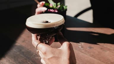 Jemand hält seine Hand an ein Espresso Martini Glas auf einem Tisch.