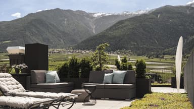 Ein Blick auf eine Terrasse mit Liegen und Sesseln und grünen Wiesen und Bergen im Hintergrund.