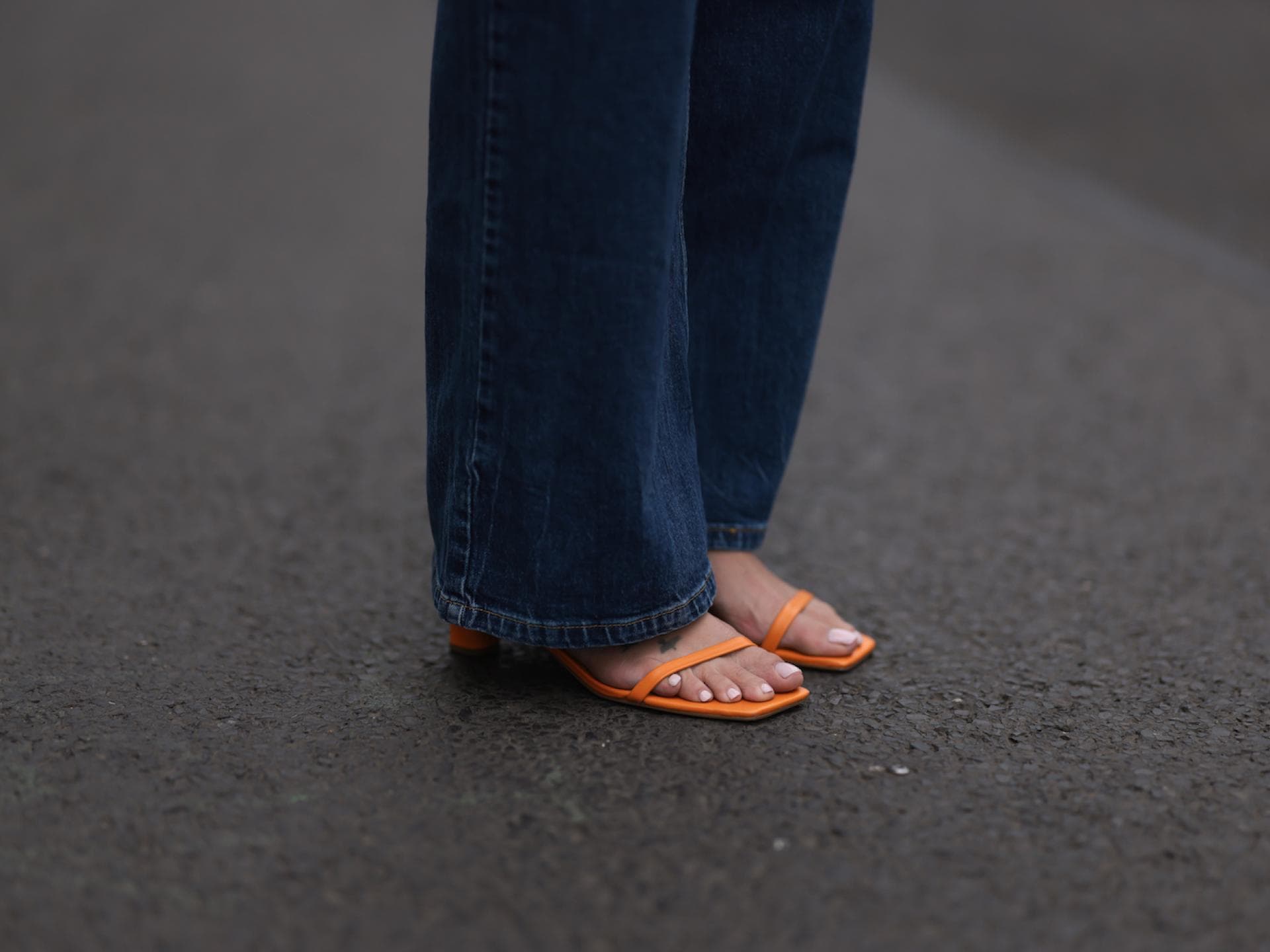 Welche Nagellackfarbe für ältere Füße geeignet ist, hängt vom Hauttyp ab. Nackte Füße in orangenen Sandalen mit hellem Nagellack und einer langen, dunklen Jeans.