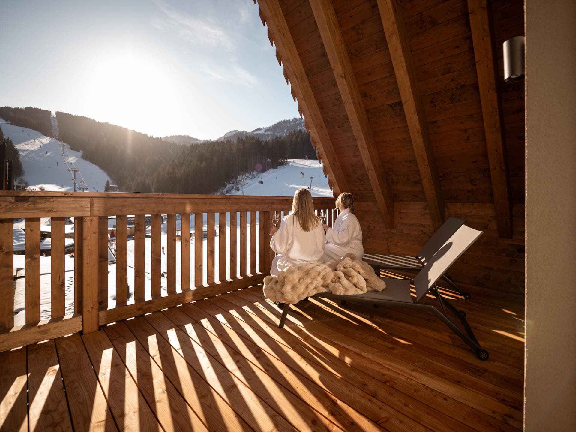 Die Adea Lifestyle Suites Fieberbrunn bieten jede Menge Erholung. Zwei Frauen sitzen auf einem Holz-Balkon mit Bademänteln und trinken Wein. Der Ausblick ist eine Landschaft in den Bergen mit Schnee.