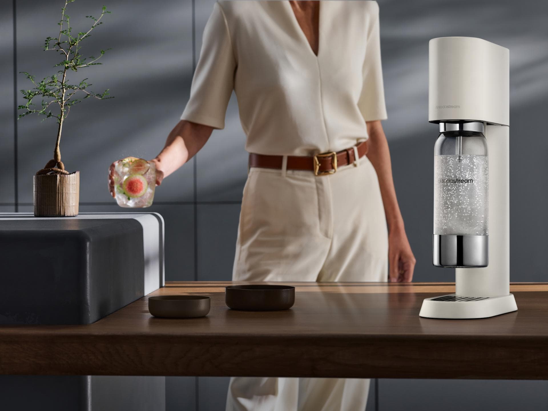 Der neue SodaStream Enso ist in einem eleganten Sand erhältlich. Eine Frau hält ein Glas mit Grapefruit Scheiben und Wasser, daneben steht ein SodaStream auf der Arbeitsfläche in der Küche.