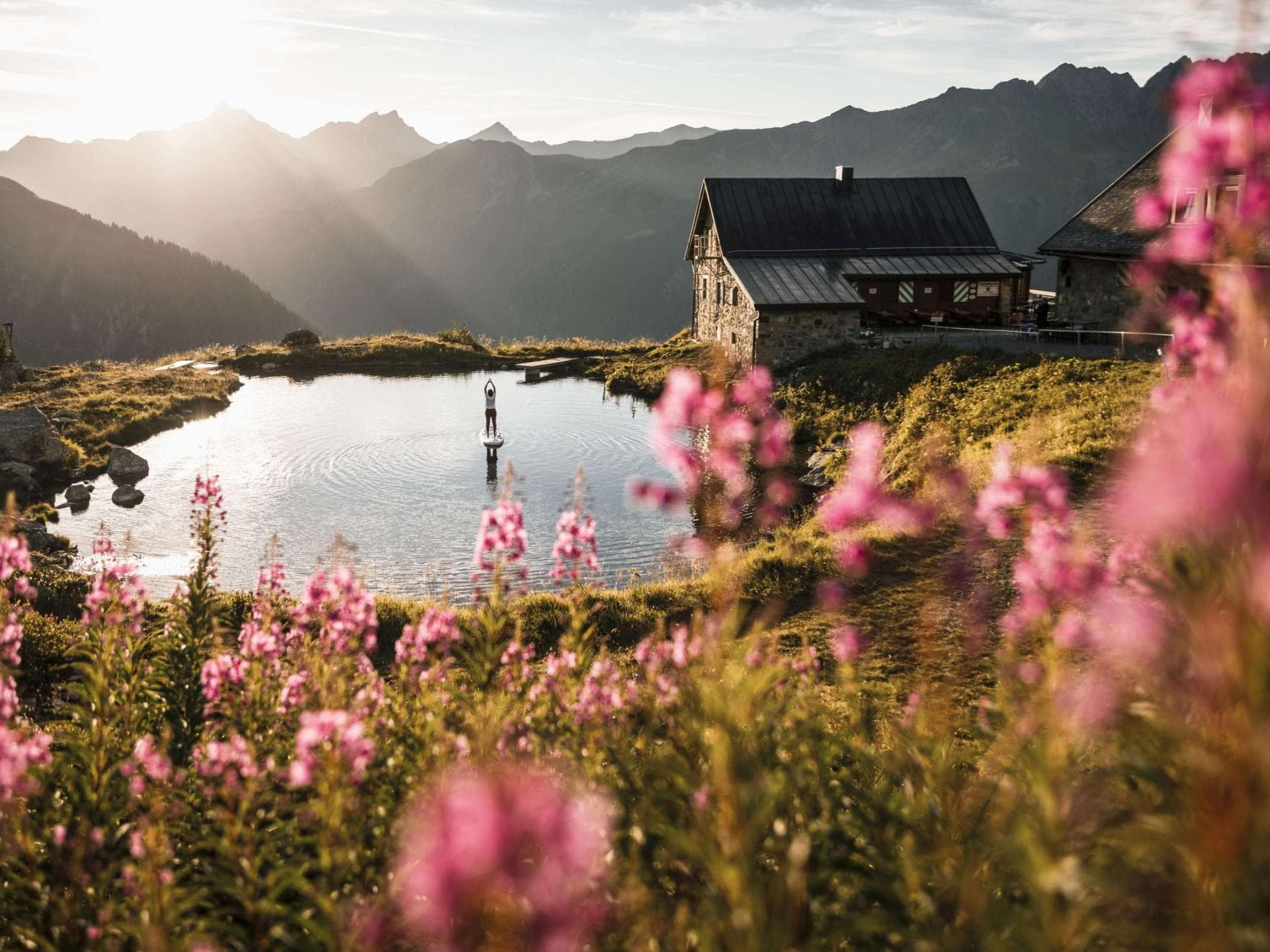 Ein Haus am See mit Gebirge im Hintergrund, rosanen Blüten im Vordergrund und Sonnenstrahlen.