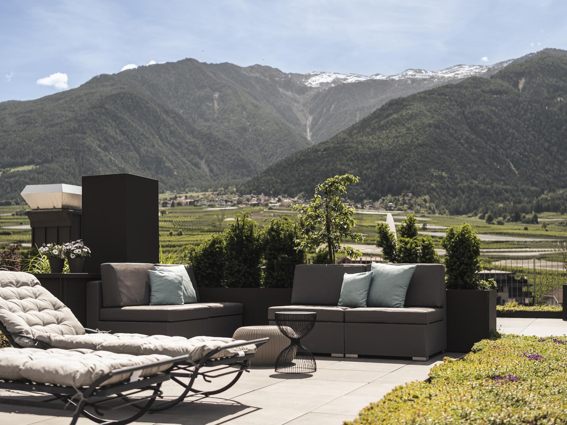 Ein Blick auf eine Terrasse mit Liegen und Sesseln und grünen Wiesen und Bergen im Hintergrund.