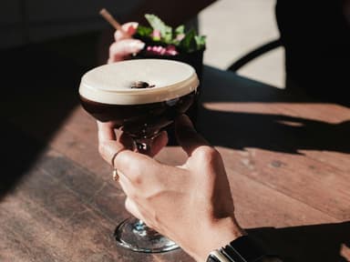 Jemand hält seine Hand an ein Espresso Martini Glas auf einem Tisch.