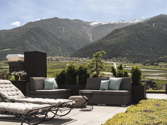Ein Blick auf eine Terrasse mit Liegen und Sesseln und grünen Wiesen und Bergen im Hintergrund.